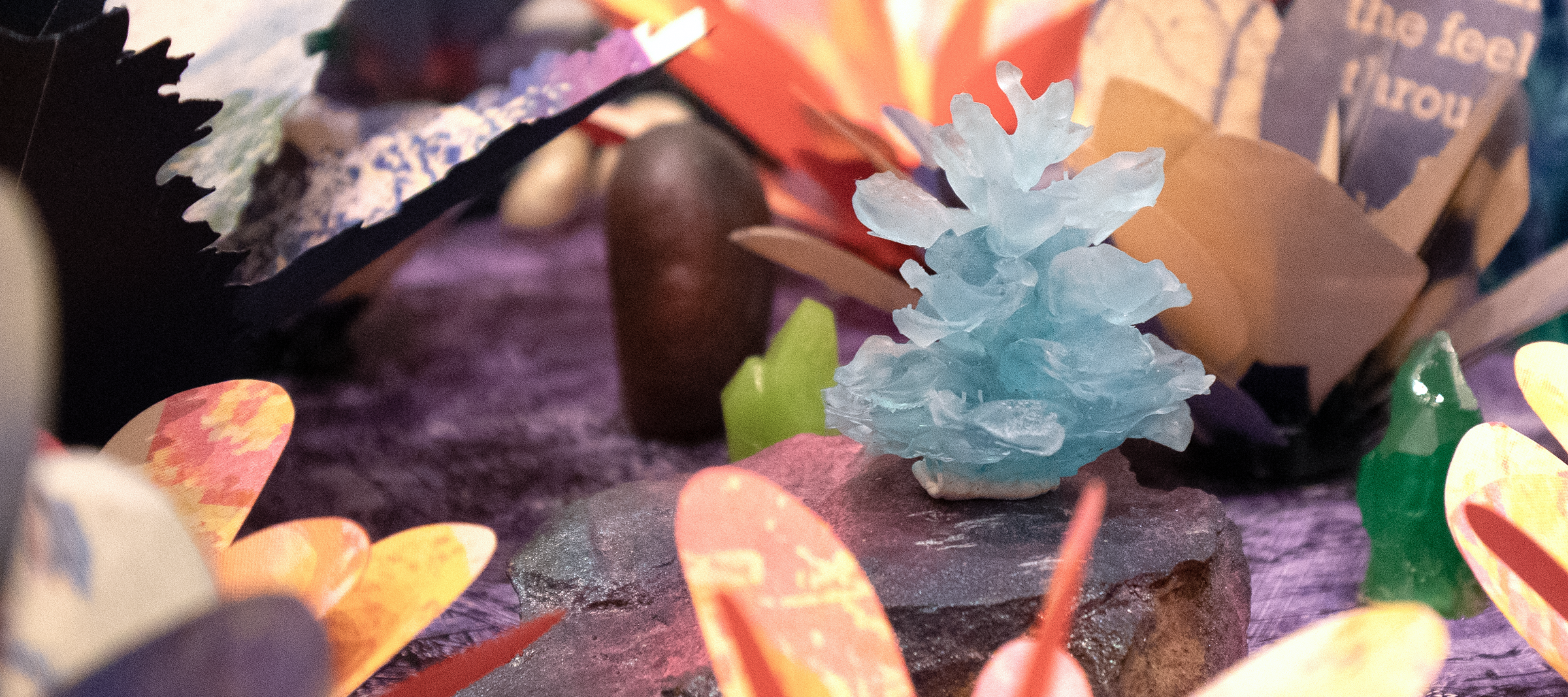 A close-up of a small, light blue crystal formation displayed on a rock, surrounded by colorful, paper-like flower shapes and decorations on a purple textured surface.