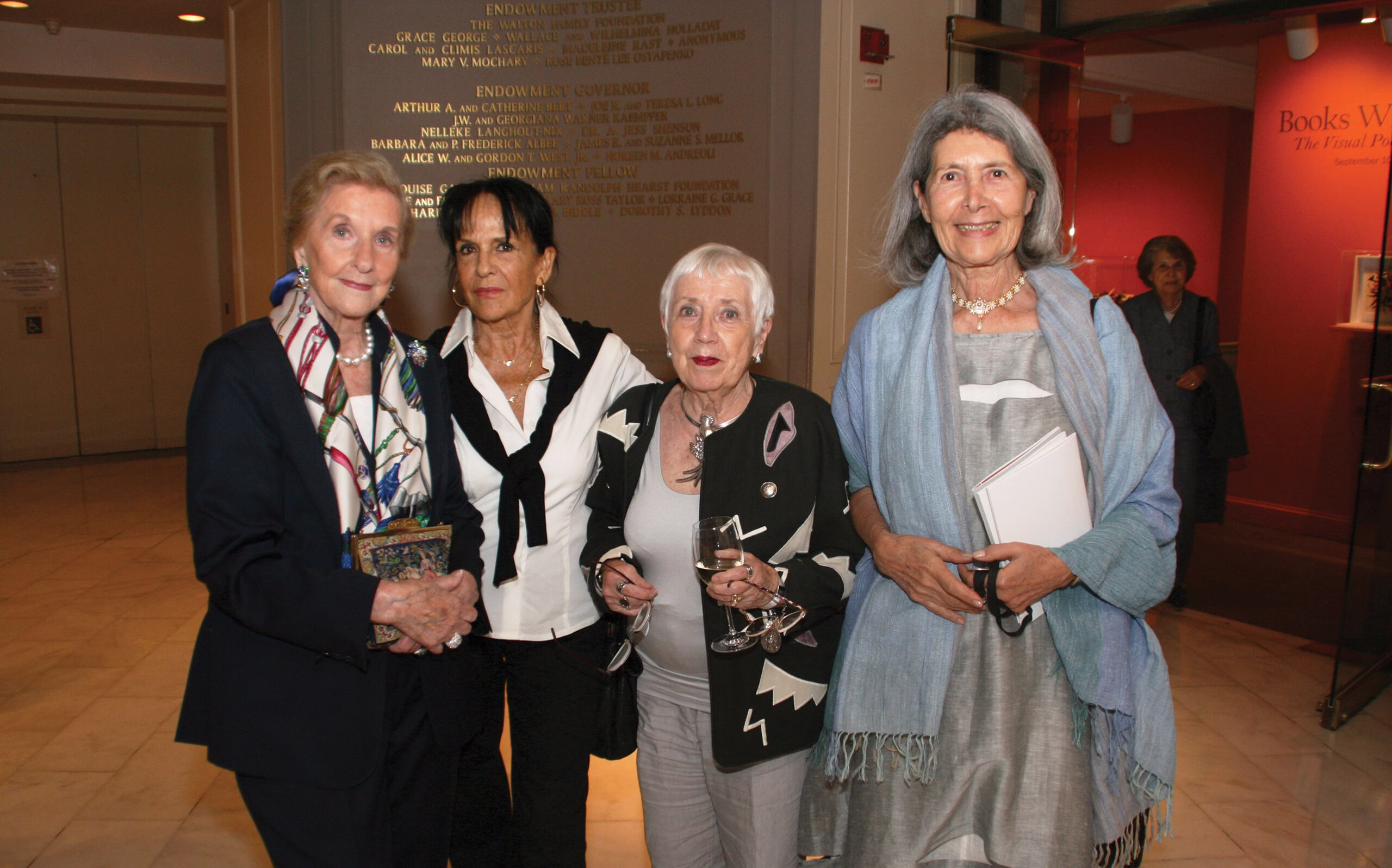 Four older women stand together at an indoor event. One holds a drink, one holds a notebook, and all are dressed formally. A wall with engraved names and a pink room are visible in the background.