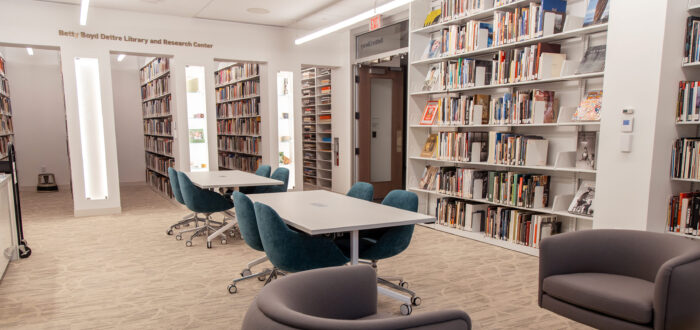 A room with shelves of books on its walls, two tables with blue chairs, and two dark grey armchairs with side tables.