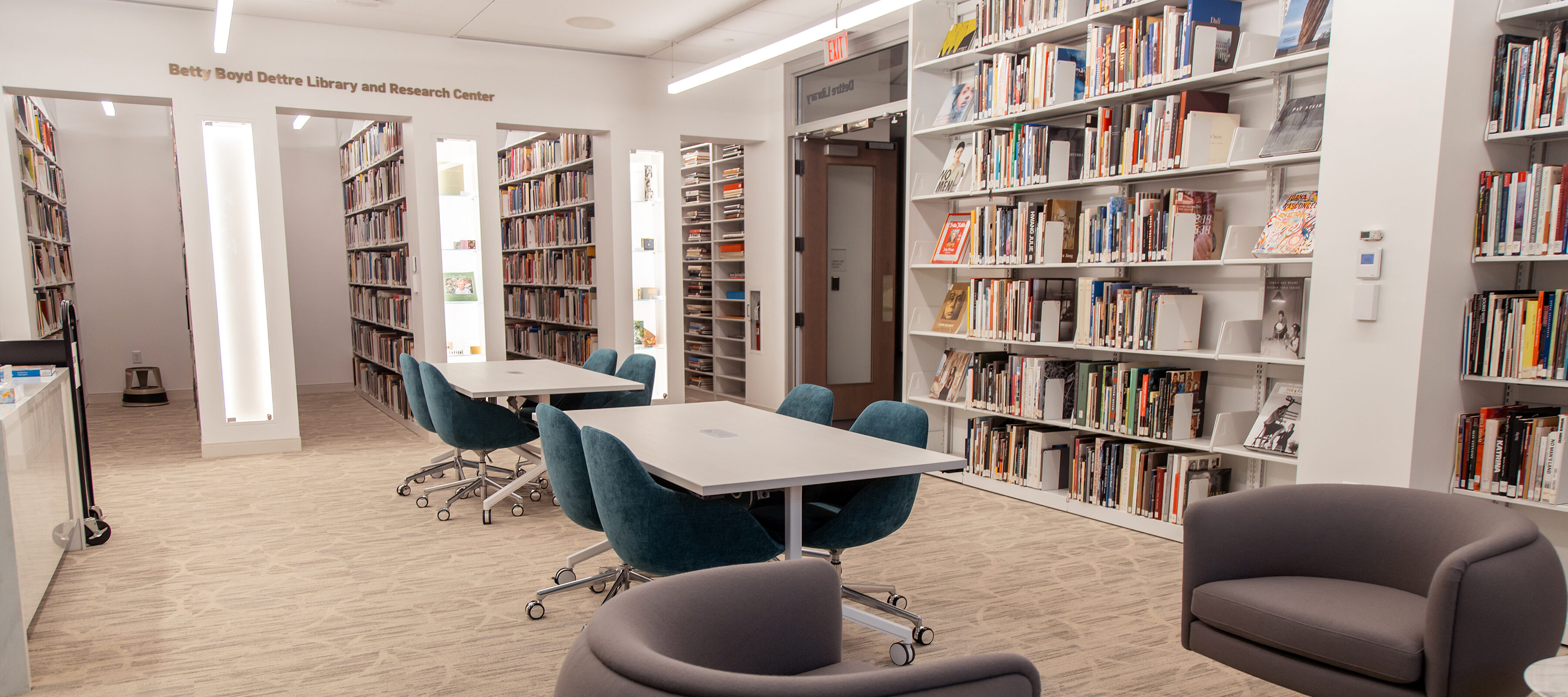 A room with shelves of books on its walls, two tables with blue chairs, and two dark grey armchairs with side tables.
