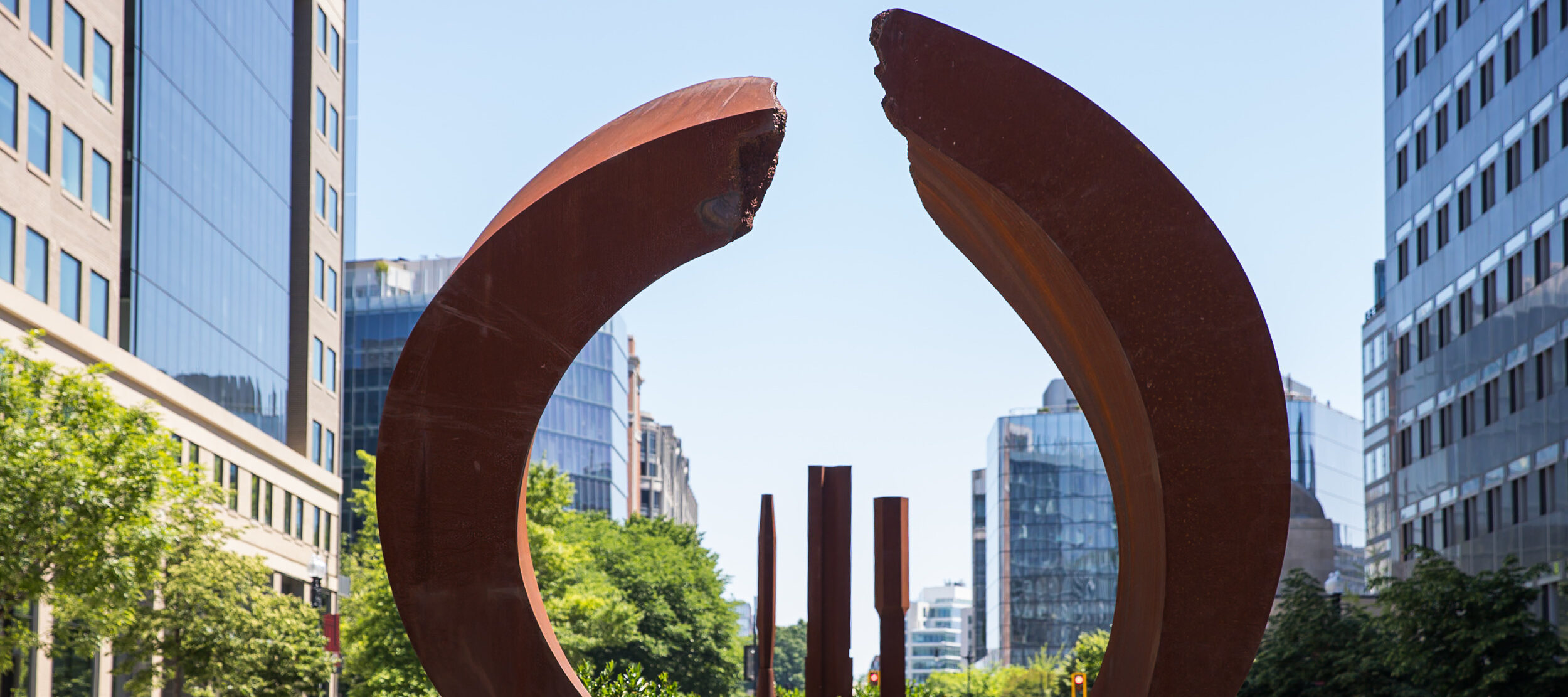 A circular, rust-colored sculpture and three rust-colored columns in the median of a city street.
