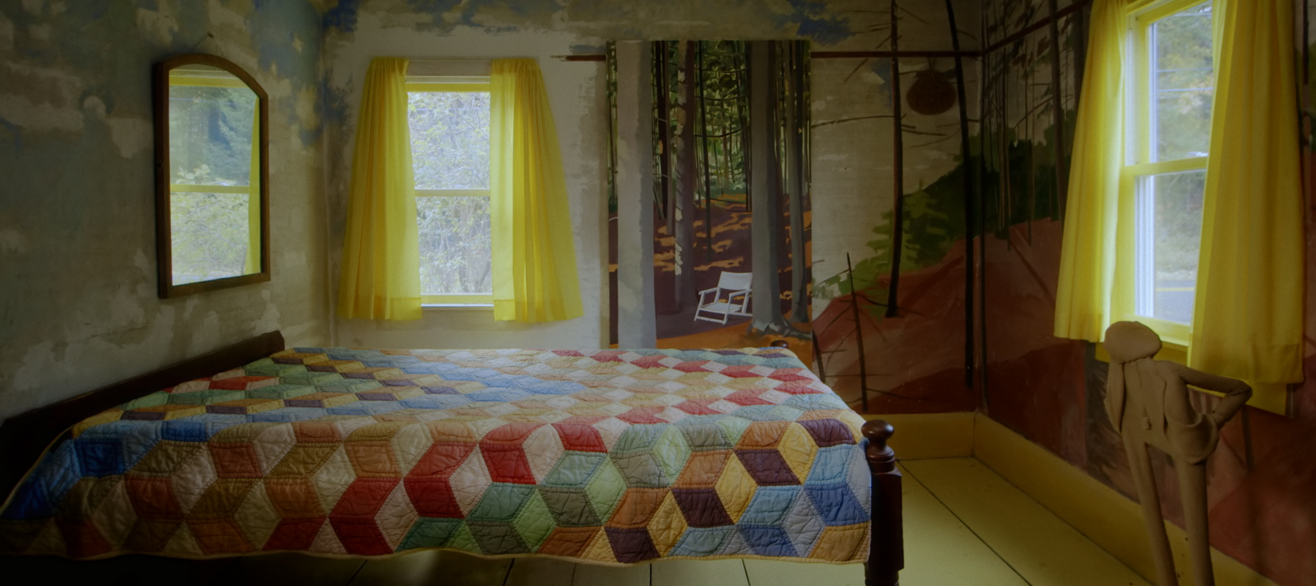 Film still of a dimly lit bedroom. The four-postered wooden bed has a multicolored quilt printed with three-dimensional cube pattern. The walls have a woodsy mural and the open light yellow curtains on the windows bring in warm light.