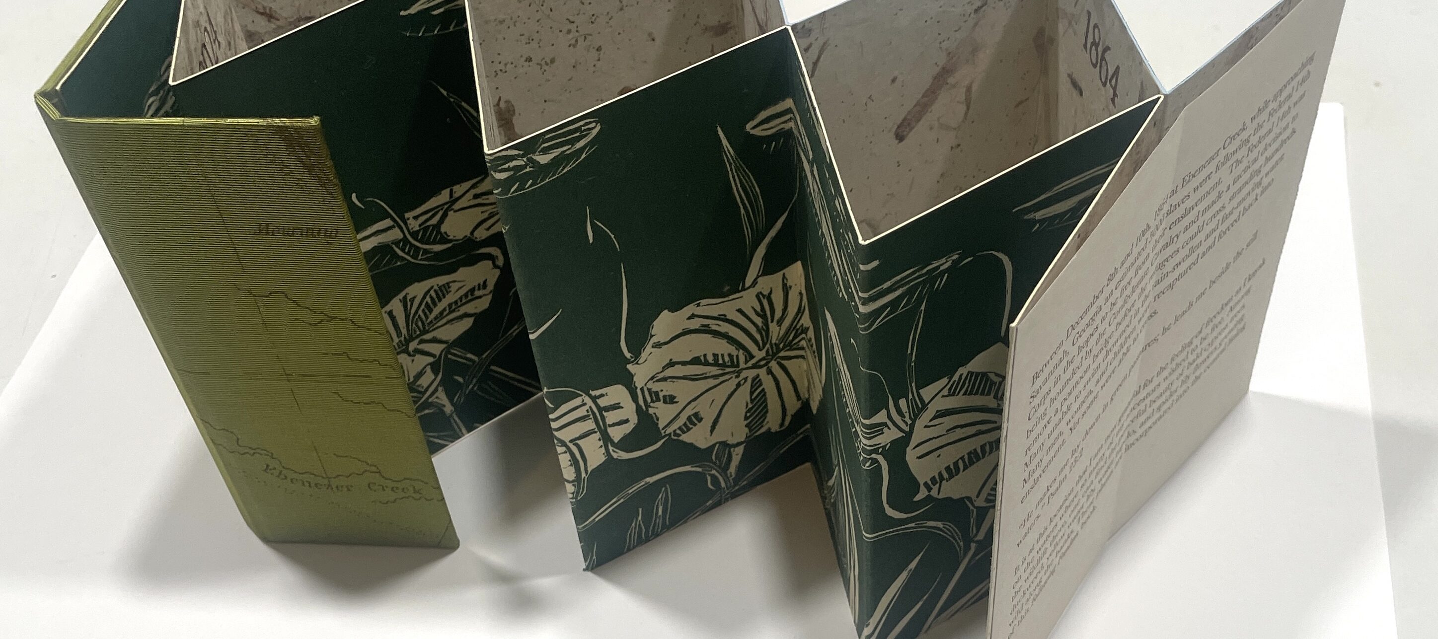 Accordion-style book entitled "Following Freedom" with green botanical prints and text is standing open on a white surface, showing patterned and text-filled pages.