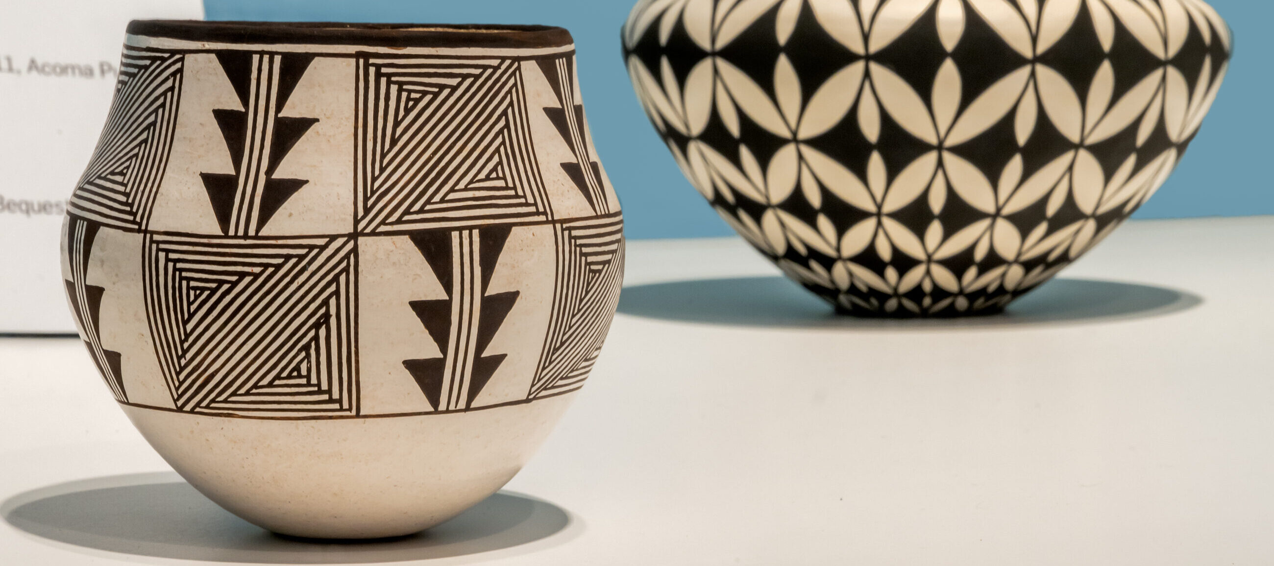 Two Pueblo ceramic vessels with black-and-white geometric designs. The piece in the foreground is round with triangular patterns and the piece in the background is a pointed vase with interlocking floral motifs.