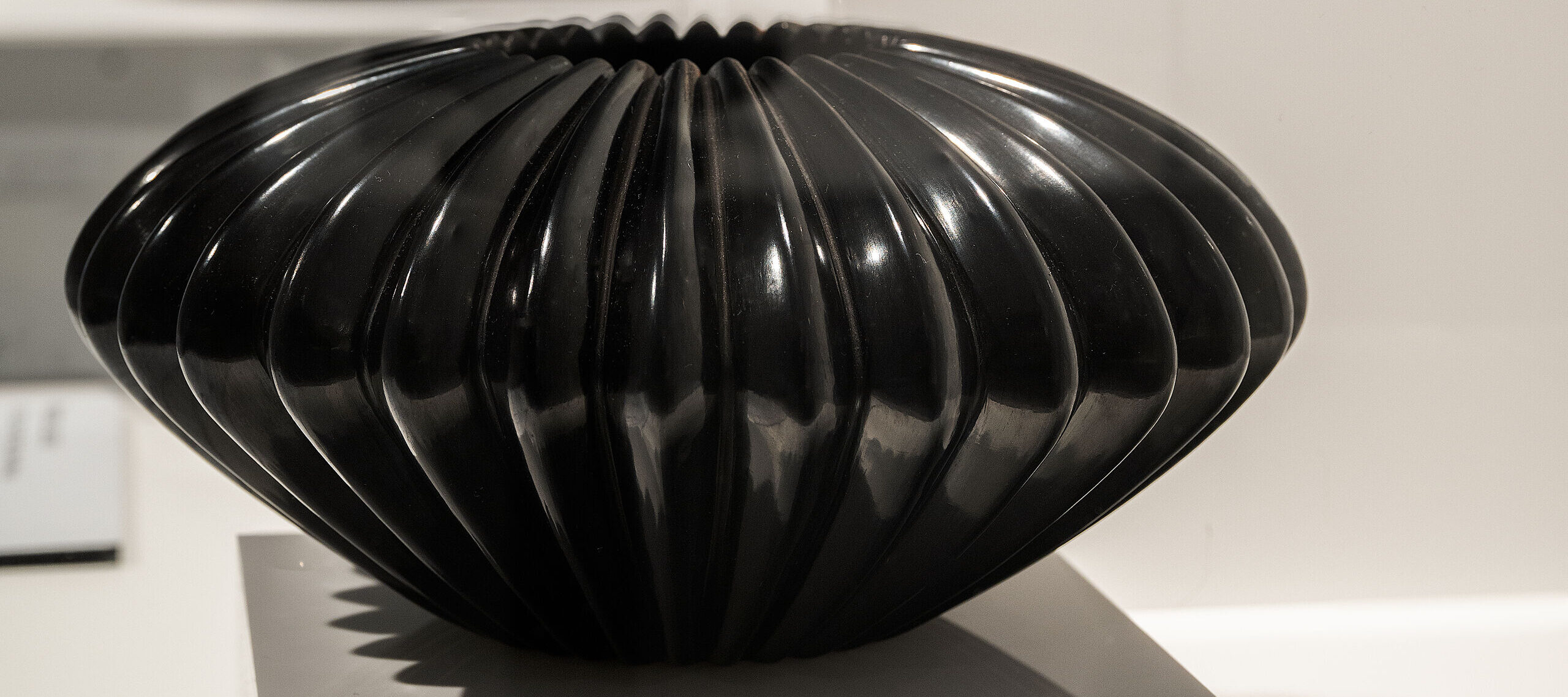 Three ceramic pots are displayed on white pedestals in a gallery setting. In the foreground is a glossy black pot with a ribbed, rounded shape. Behind it stands a vessel with a geometric black-and-white pattern and orange base. The third vessel is tall and black.