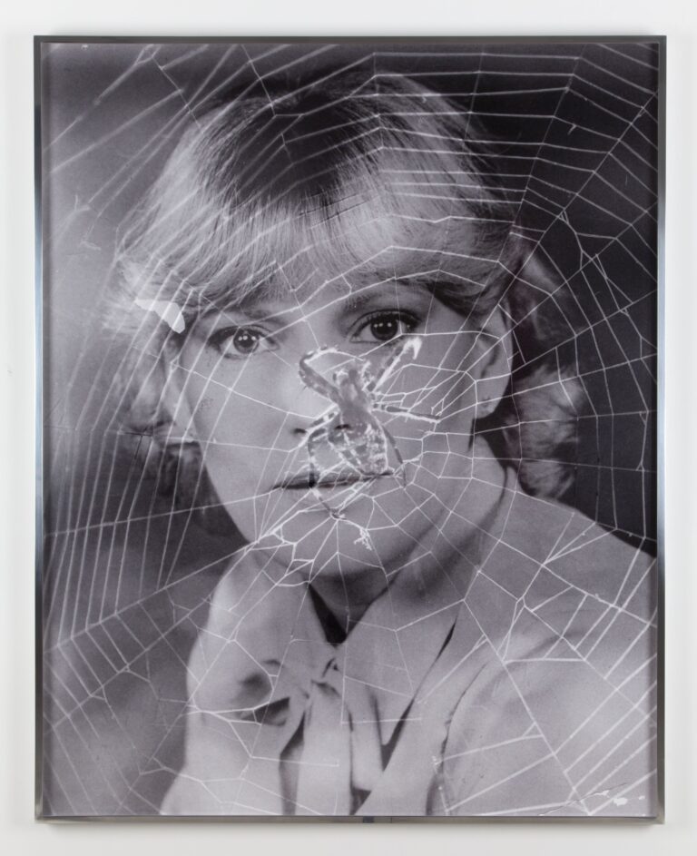 A black-and-white portrait photograph of a woman with light skin tone and light hair is overlaid with the image of a spiderweb. The web’s lines cross over her face and upper body as she gazes directly at the camera.