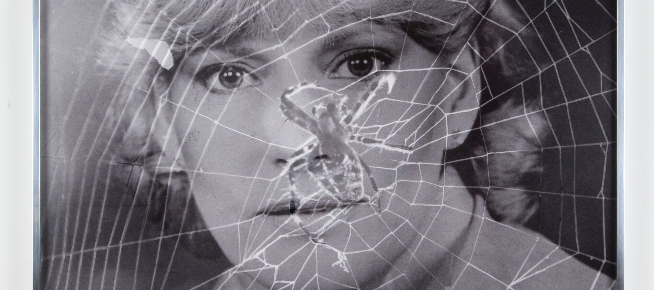 A black-and-white portrait photograph of a woman with light skin tone and light hair is overlaid with the image of a spiderweb. The web’s lines cross over her face and upper body as she gazes directly at the camera.