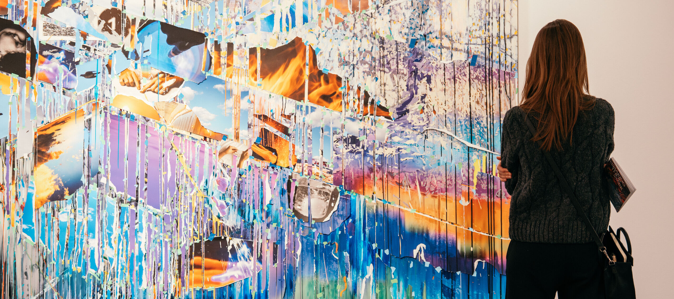 A person with long hair stands in front of a large, colorful abstract painting in an art museum, holding a small catalogue.