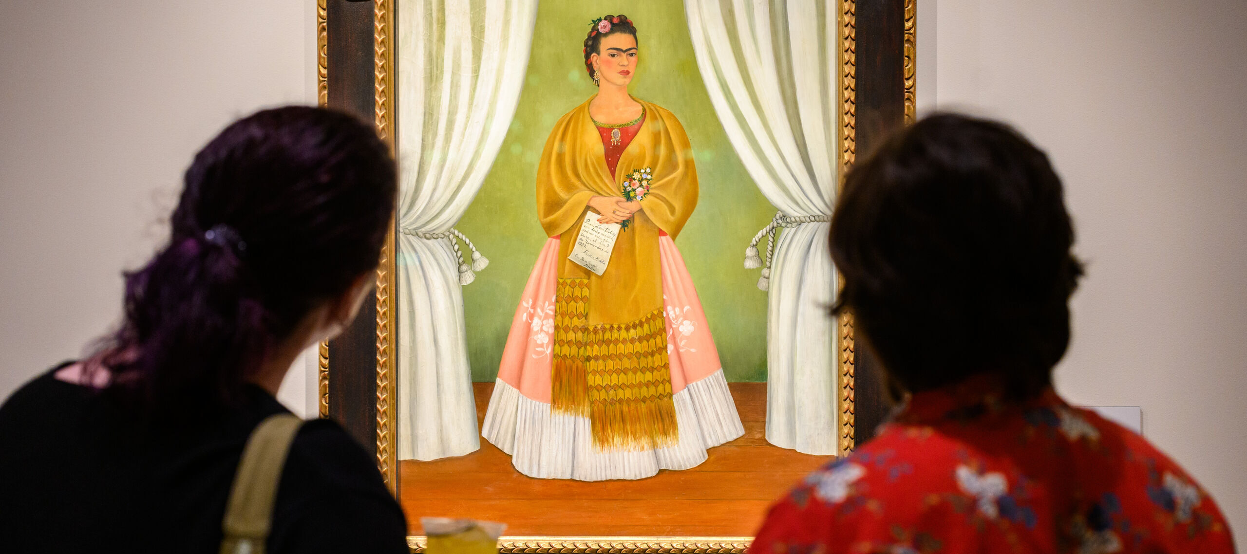 Two look at a colorful painting featuring a woman in a long pink and white skirt with a gold shawl around her shoulders.