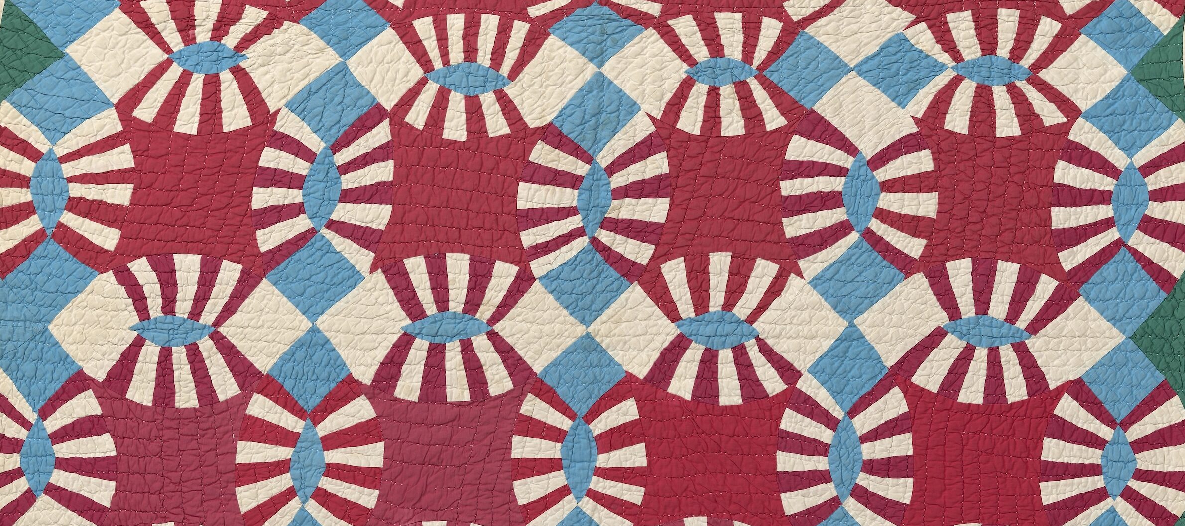 Quilt in a Double Wedding Ring pattern with interlocking rings of maroon and cream striped arcs, joined by pale blue oval lenses and maroon diamond patches, with green triangular corner blocks.