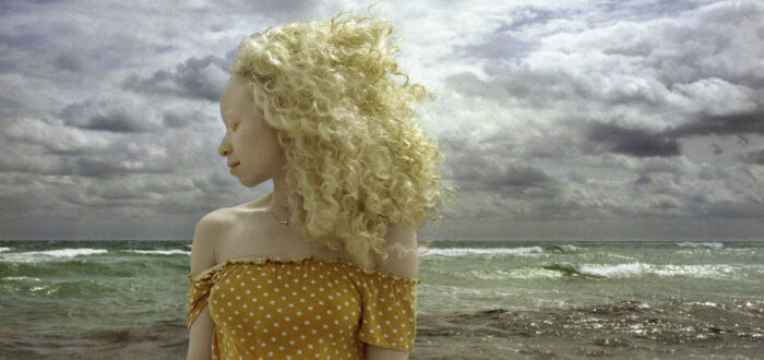 In front of a stormy ocean, a woman with light skin and blonde, wind-blown hair stands with head turned in profile and eyes closed.
