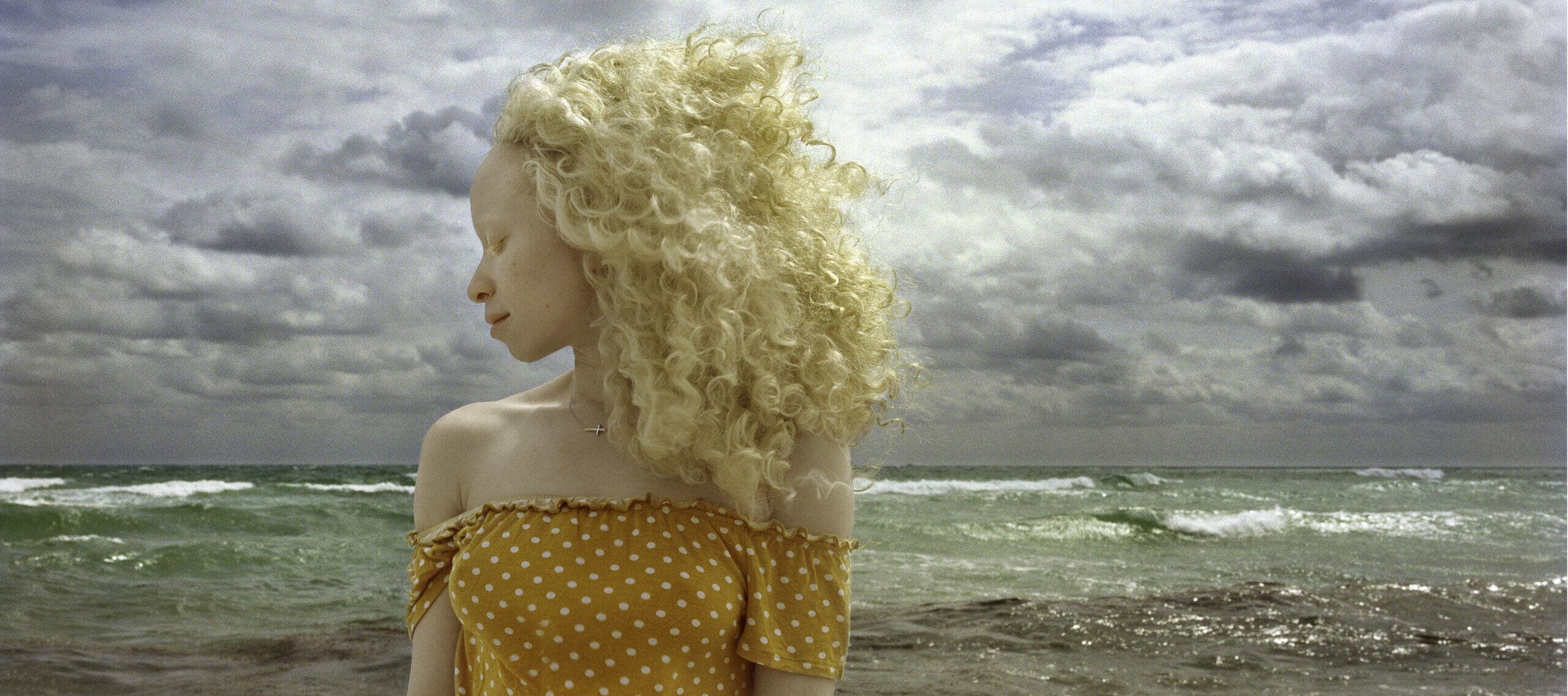In front of a stormy ocean, a woman with light skin and blonde, wind-blown hair stands with head turned in profile and eyes closed.