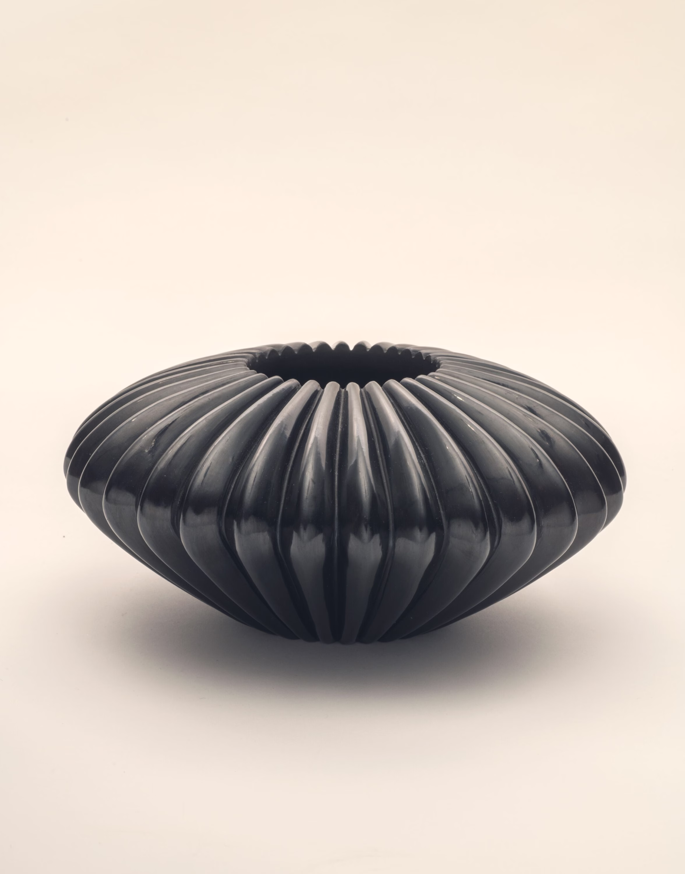A shiny, blackware vessel with deep, equidistant grooves radiating from the center opening at the vessel’s top all the way down to its base. The vessel is squat, short and very wide, resembling a squat sphere.