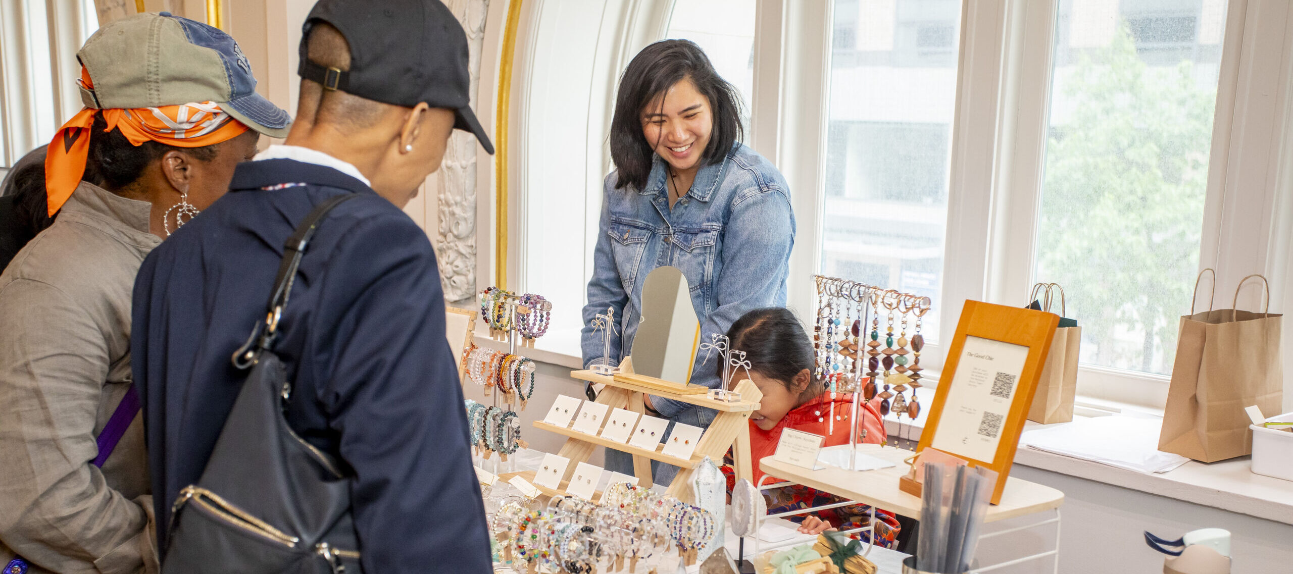 A smiling vendor stands behind a table displaying colorful handmade jewelry, while three shoppers browse the items at an indoor market with large windows in the background.