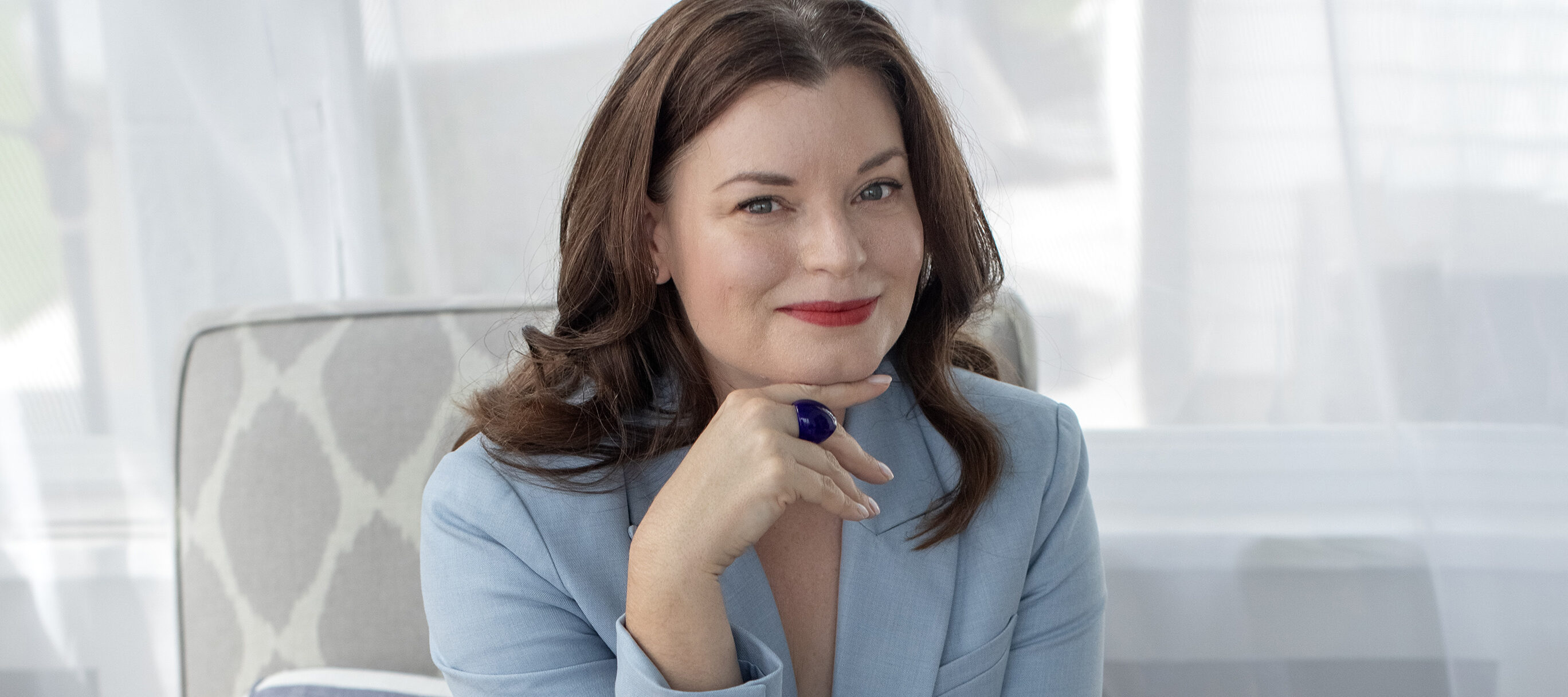 A color photograph of a woman with light skin tone and reddish-brown hair wearing a light blue suit and gold strappy sandals. She is holding a dark blue book while sitting on a chair with a gray-and-white cushioned chair.
