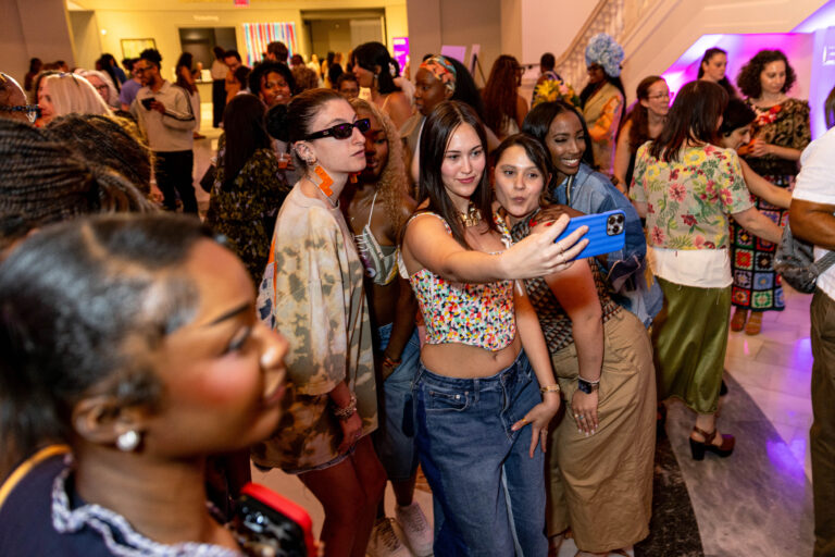 A group of young women take a selfie at a crowded indoor event. People in colorful, trendy outfits mingle and chat in the background. The atmosphere is lively and festive.
