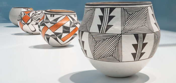 Four ceramic pots with intricate black, white, and red geometric patterns sit in a row on a white surface against a blue background. The pots feature various designs.