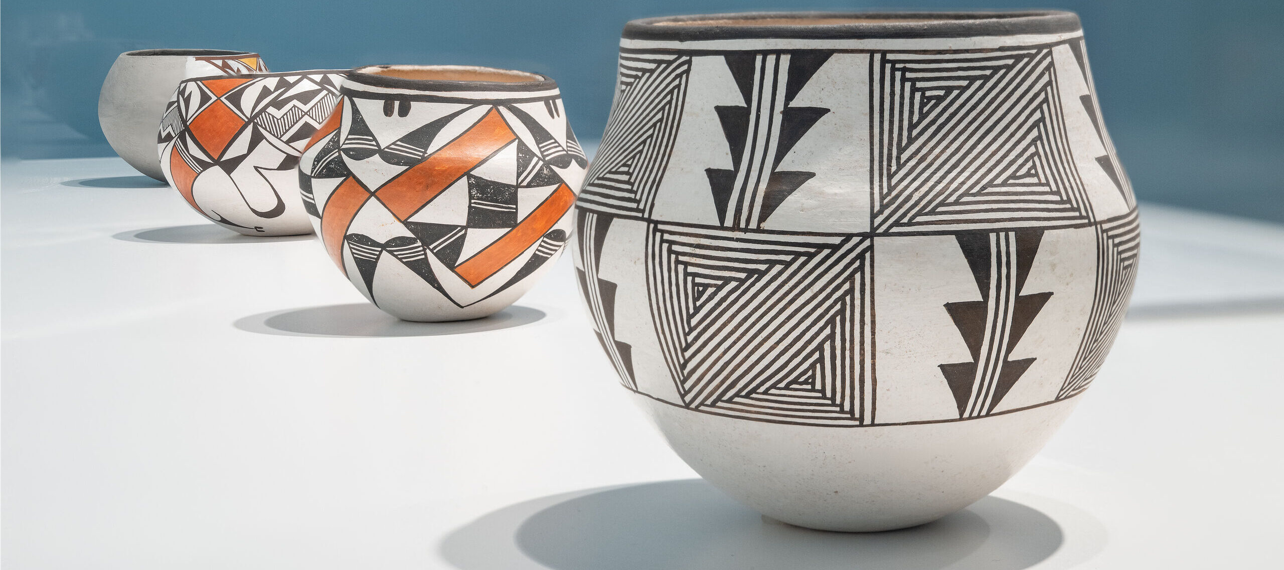 Four ceramic pots with intricate black, white, and red geometric patterns sit in a row on a white surface against a blue background. The pots feature various designs.