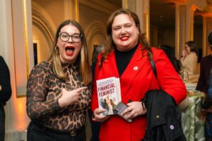 Two women smile at an event; one in a leopard print top excitedly points at the other, who is wearing a bright red blazer and holding the book "Financial Feminist." Ornate, gold-accented room in the background.