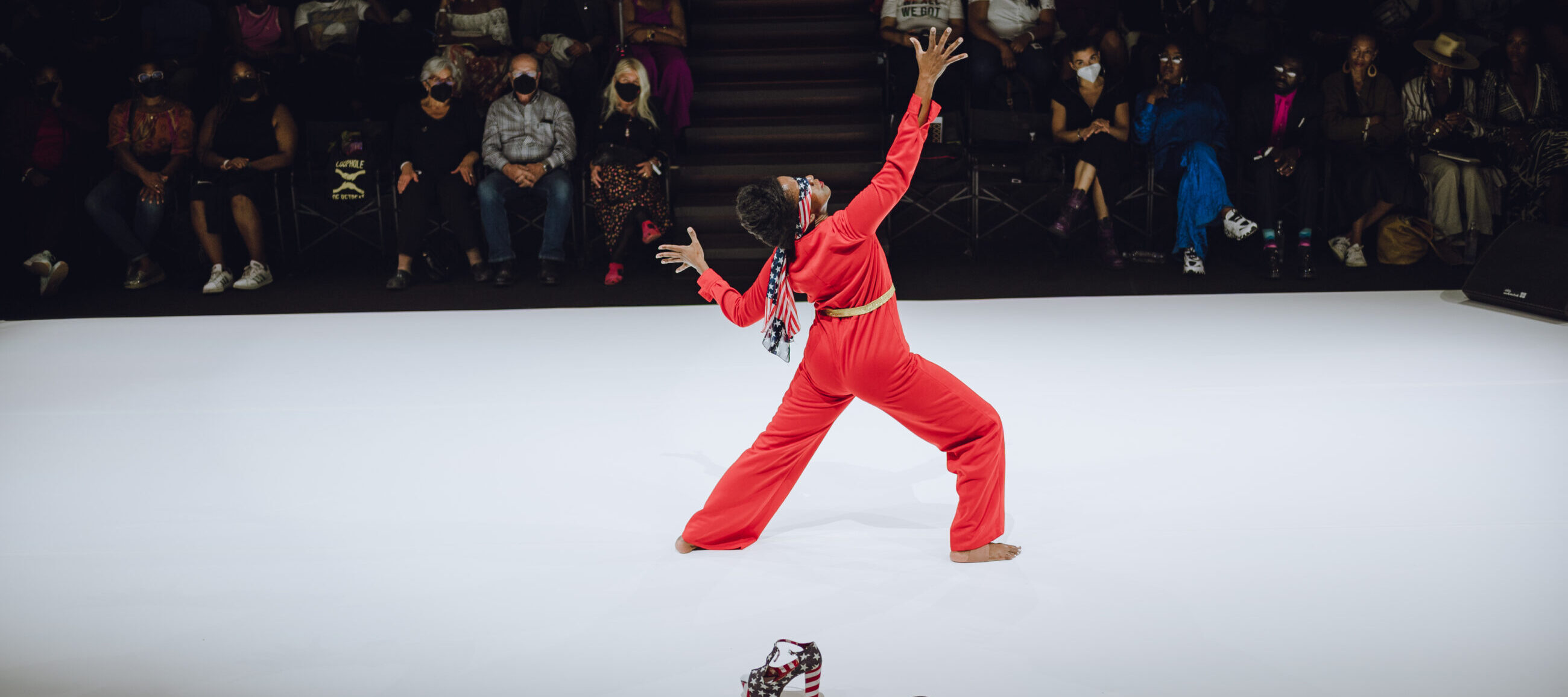 A performer in a red outfit poses barefoot on a white runway, with patterned high-heeled shoes placed in front; audience members watch in the background.