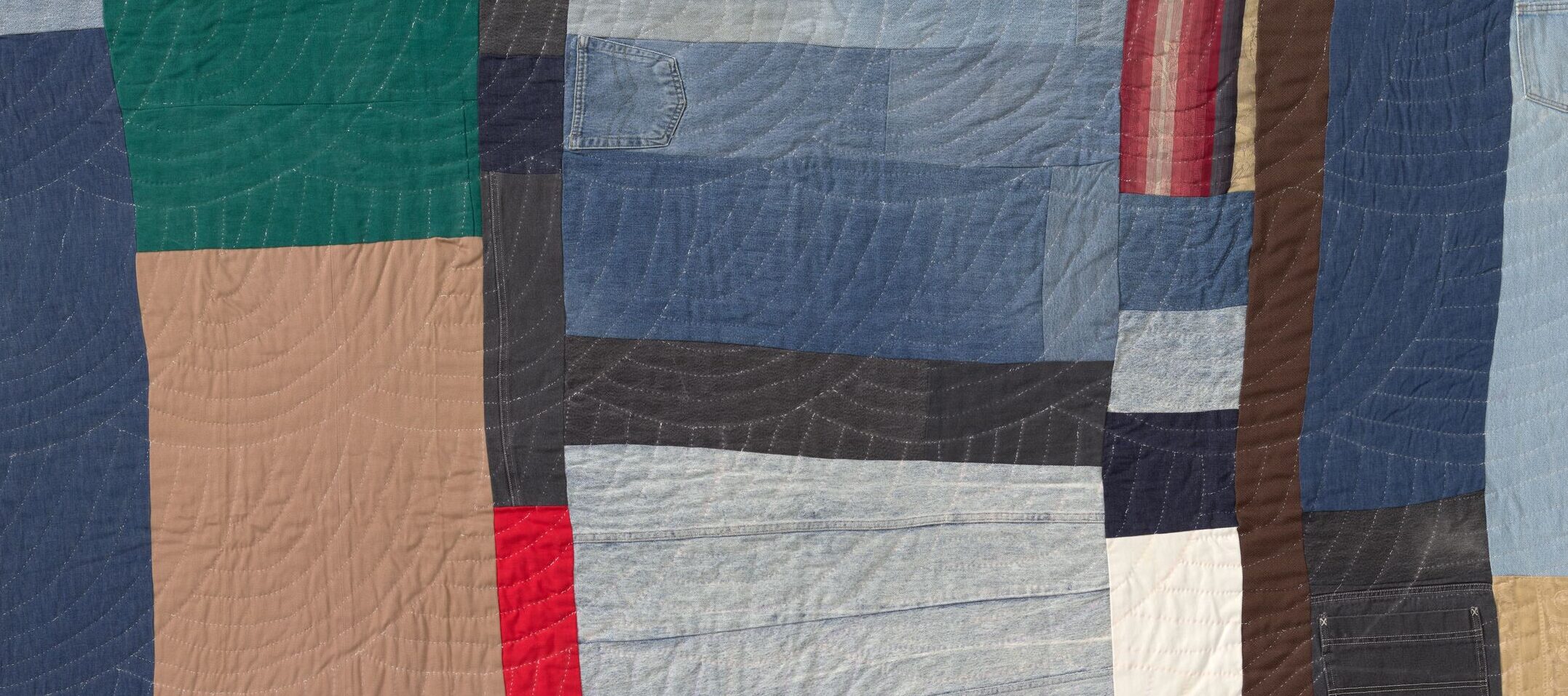 A quilt made from uneven, colorful fabric strips. There are blocks of blue, tan, gray, black, red, and green arranged in an abstract, patchwork pattern.