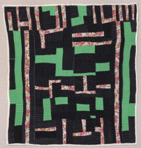 A quilt with geometric shapes comprised of irregular scraps of black, green, and a red floral print fabric.