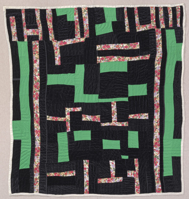 A quilt with geometric shapes comprised of irregular scraps of black, green, and a red floral print fabric.