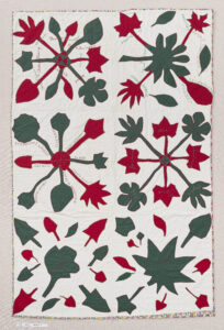 A white quilt with green leaves and red floral shapes appliquéd to the surface. Text in black stitches encircles the shapes.