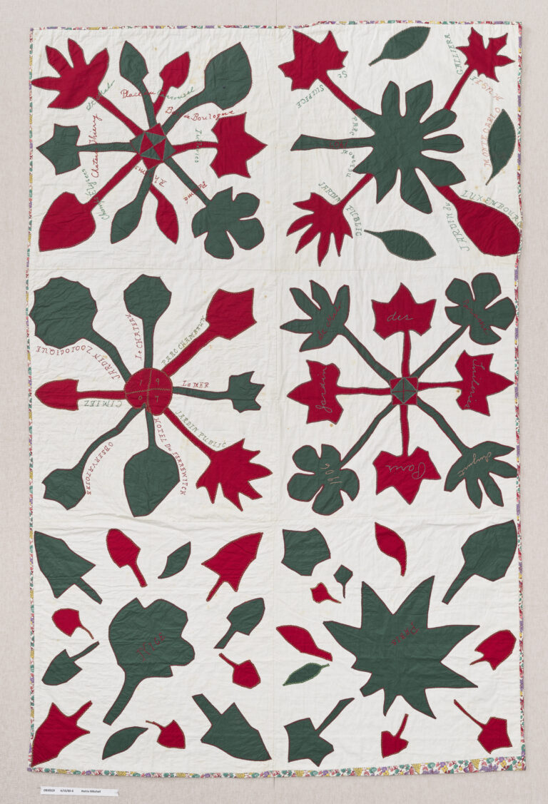 A white quilt with green leaves and red floral shapes appliquéd to the surface. Text in black stitches encircles the shapes.