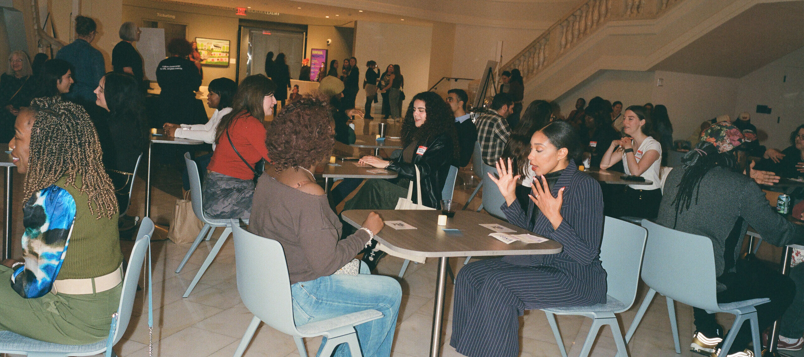 People sit at small tables in a spacious, elegant hall with a white marble floor and a balcony above. Many are engaged in conversation while others watch from the upper level. The atmosphere is lively and social.