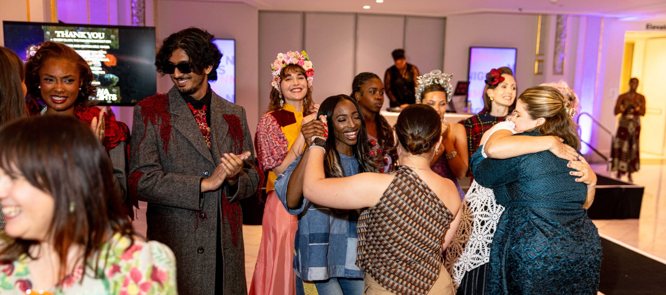 A group of people, some in colorful costumes and floral crowns, are smiling, hugging, and celebrating in a brightly lit indoor venue with a stage in the background.