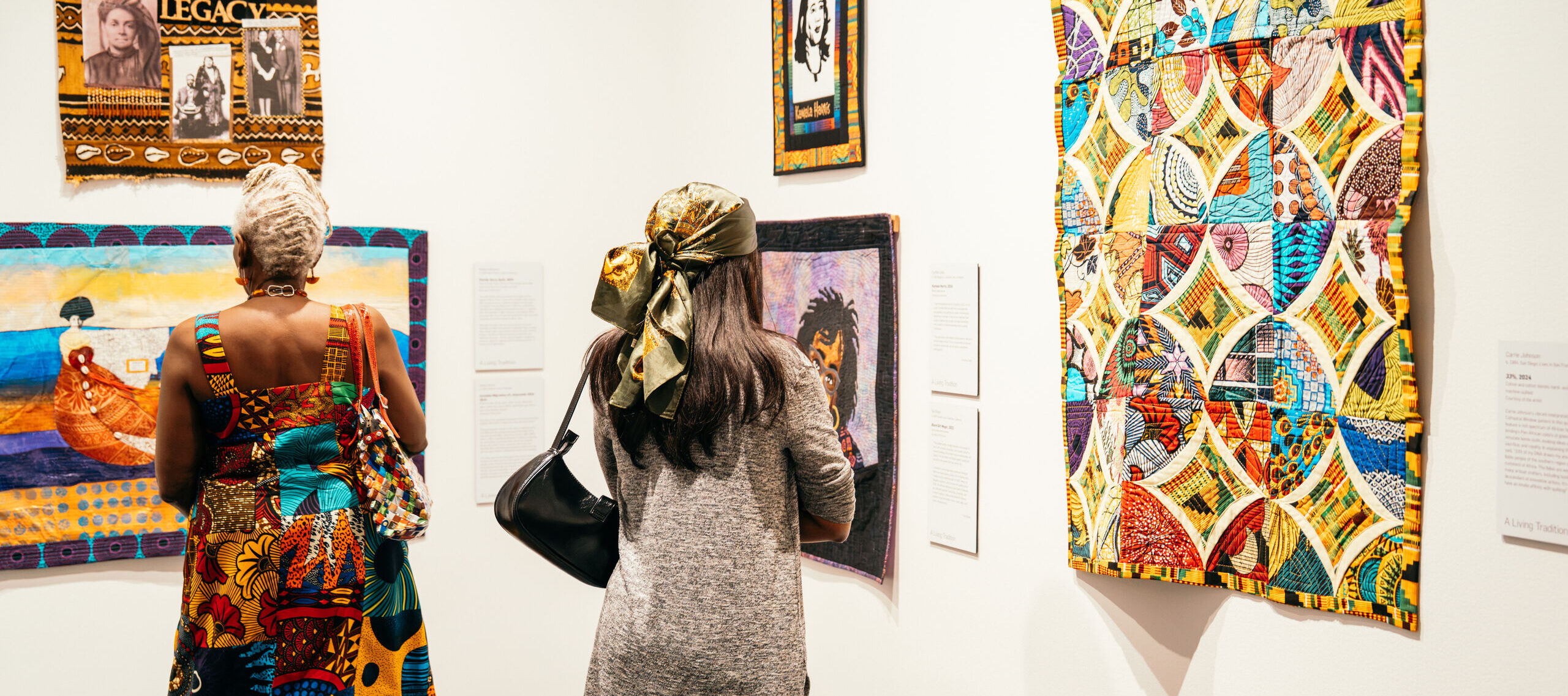Two people view colorful quilts and framed art on a white gallery wall. One visitor wears a patchwork dress, the other a gray sweater and scarf. The art features vibrant patterns and portraits.