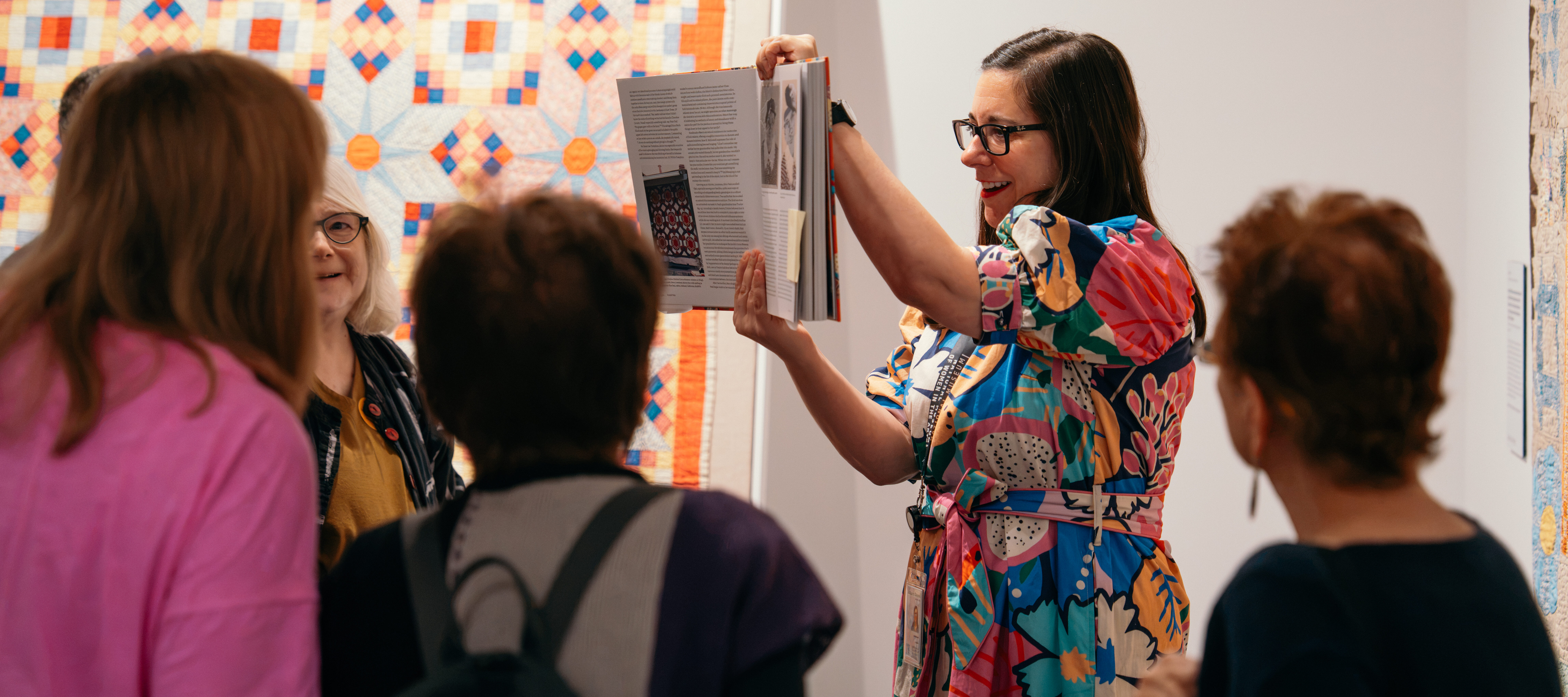 A woman with dark brown hair wearing a colorful dress and dark colored glasses holds a book to a group of visitors in front of two multicolored quilts.